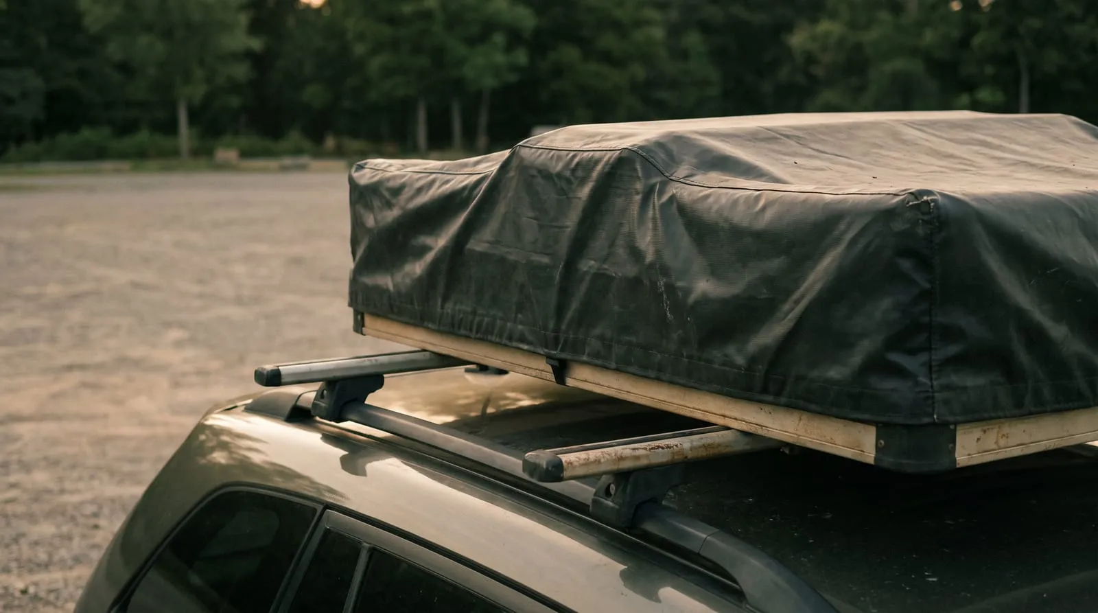 Ein Dachzelt unter einer schwarzen Transporthaube auf den Querträgern eines geparkten Kombis, dahinter eine Schotterfläche und Wald